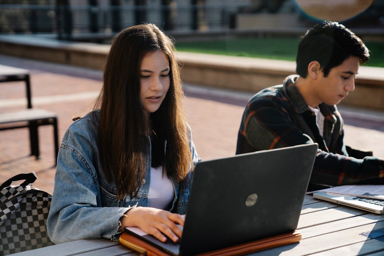 Two young adults working on laptops at an outdoor table.